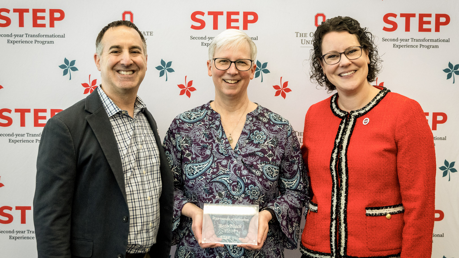 Three people smiling into the camera while standing in front of a backdrop with the person in the middle holding an award
